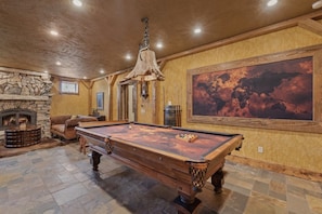 A pool table in a room with a stone fireplace, wooden walls, and a large painting.
