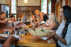 A group of people dining at a wooden table with a rustic interior, including a large window and a kitchen area in the background.