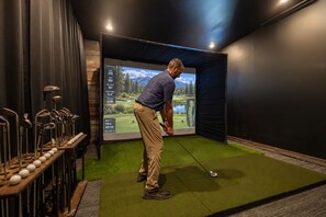 Een man oefent golf in een ruimte met kunstgras, omringd door golfclubs en een groot scherm waarop een golfbaan wordt weergegeven.