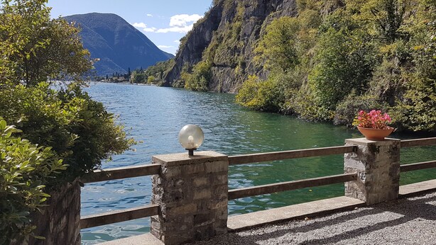 A stone bridge with a metal railing, a flower pot, and a clear view of a lake surrounded by mountains. - Directly on Lago di Lugano with boat mooring (Porlezza)