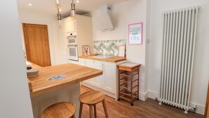 Una cocina moderna con encimeras de madera, horno empotrado y radiador blanco. Hay dos taburetes y un pequeño carrito de madera.