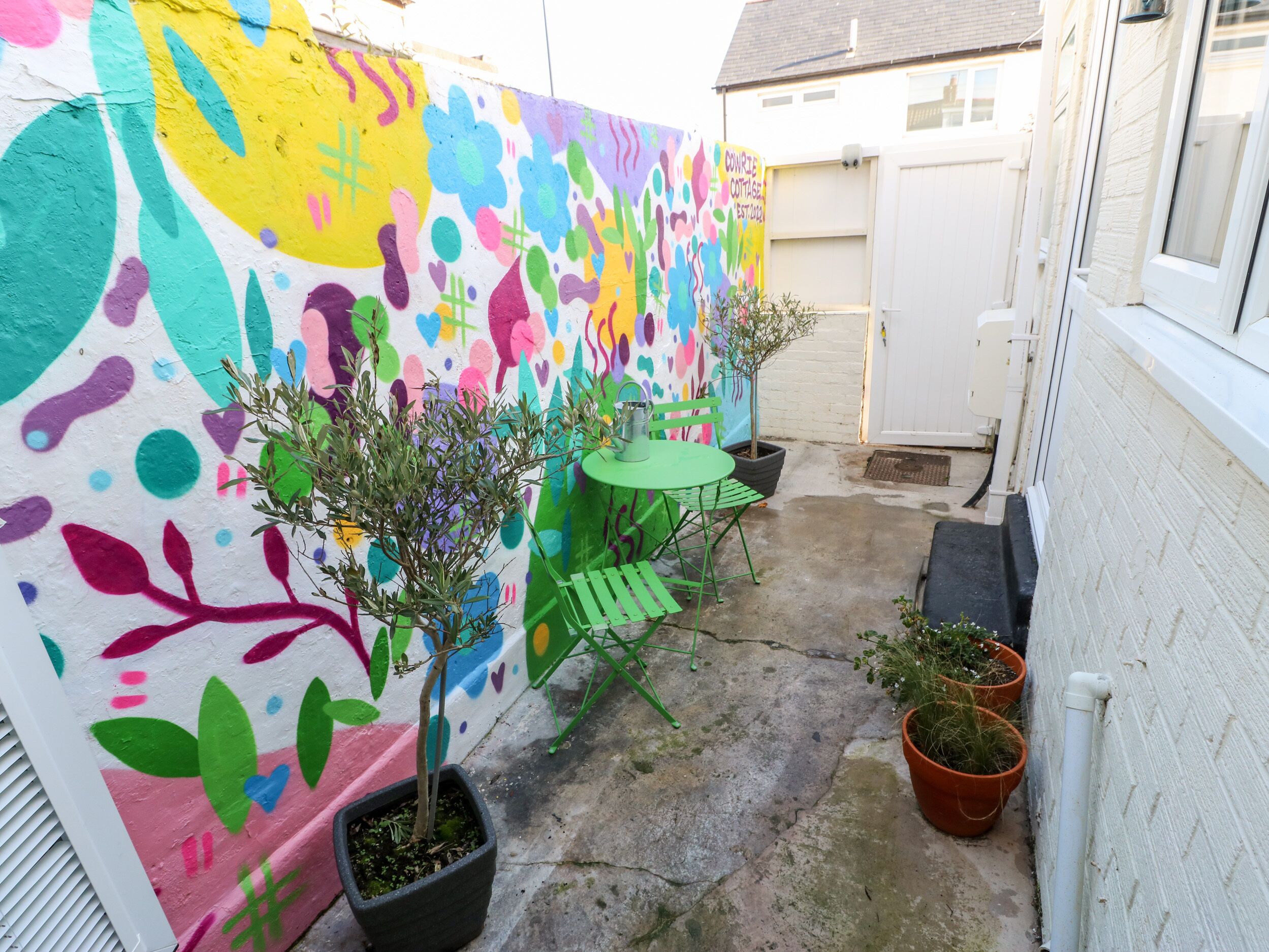 Un pequeño patio al aire libre con un mural colorido, una mesa verde y plantas en macetas.