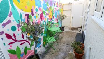 Un pequeño patio al aire libre con un mural colorido, una mesa verde y plantas en macetas.