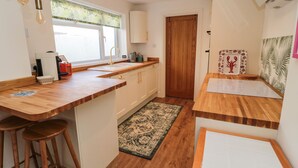 Cocina compacta con encimeras de madera, un lavamanos y una ventana con cortina floral. Hay taburetes, una alfombra y una pequeña mesa con un objeto decorativo.