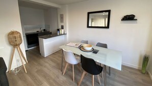 Un espace repas moderne avec une table en verre, deux chaises blanches et une chaise noire. On aperçoit, au fond, un petit îlot avec un évier et un plan de travail.