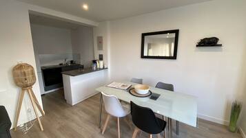 A modern dining area with a glass table, two white chairs, and a black chair. A small island with a sink and counter is visible in the background.