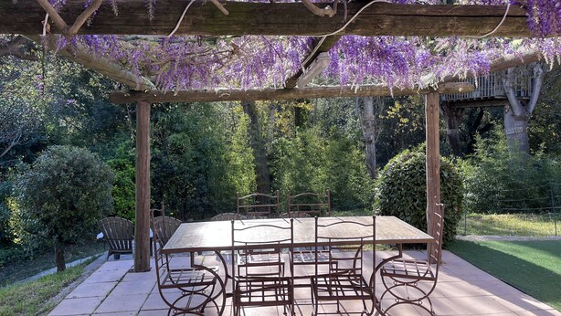 Une terrasse couverte, agrémentée d’une table et de chaises, entourée d’une végétation luxuriante et de plantes grimpantes aux fleurs violettes.
