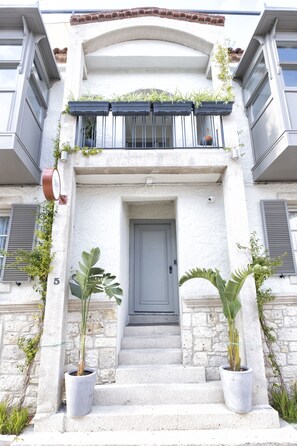 A building with a white facade, a grey door, and steps leading to a balcony with potted plants. - Alle Alacati (Cesme)