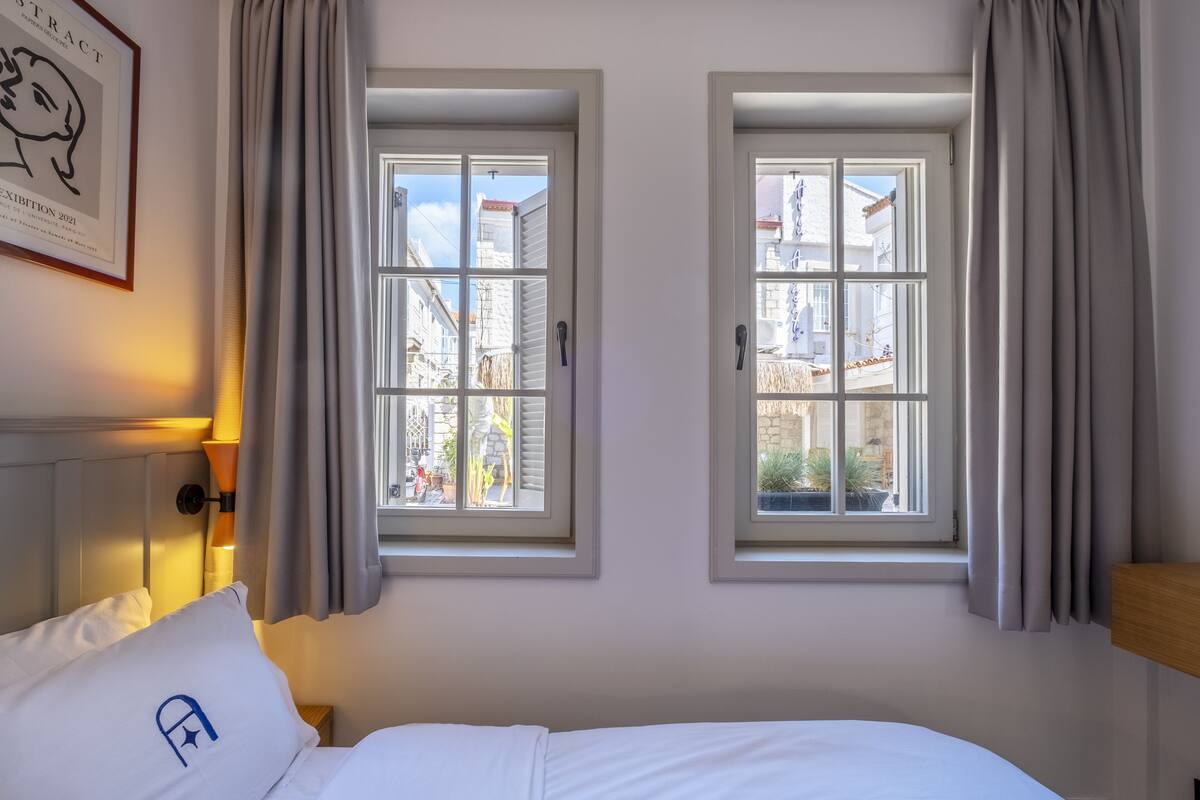 A hotel room with two windows, a bed with white bedding, and a framed picture on the wall.