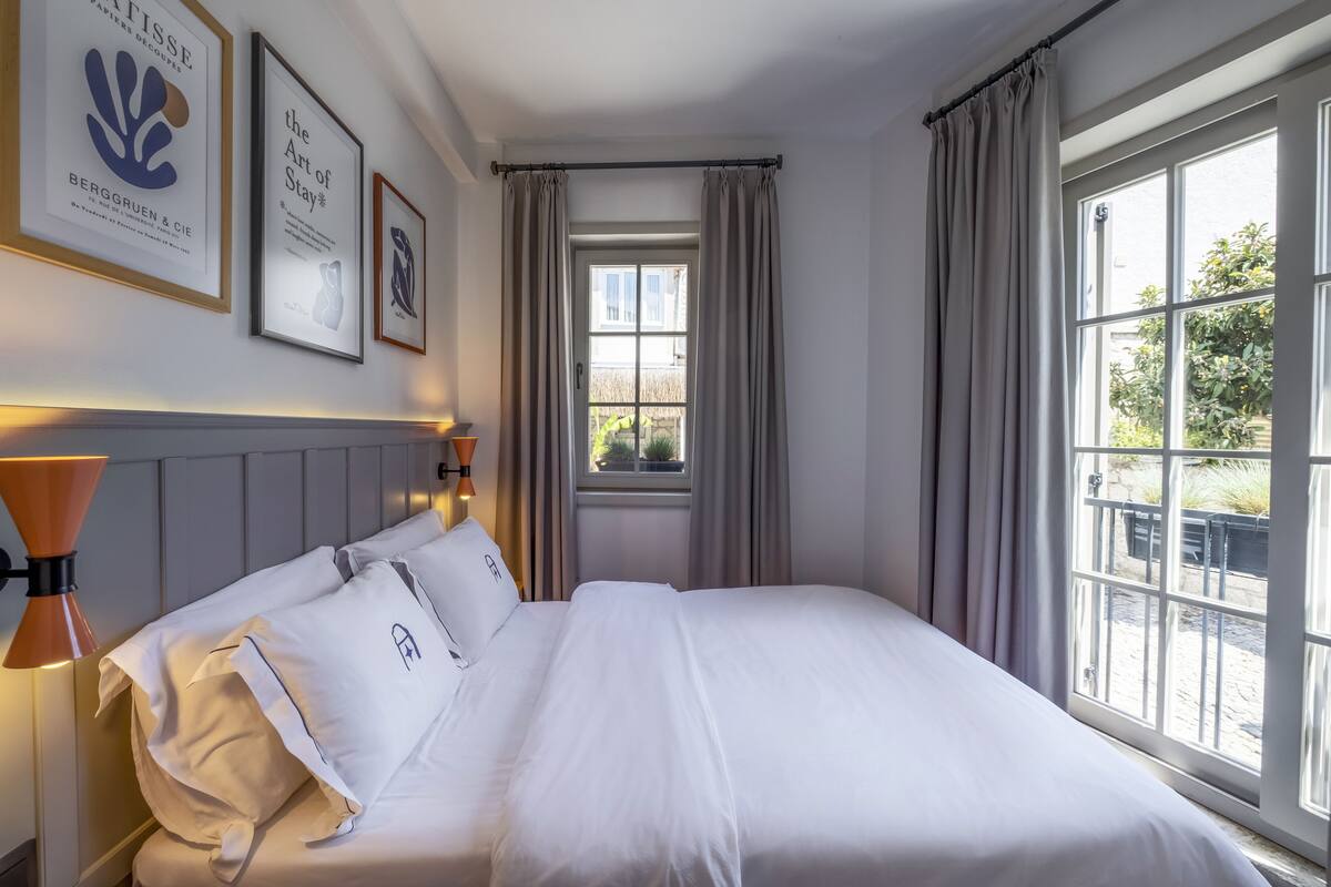 A neatly made bed in a hotel room with a large window, curtains, and framed artwork on the wall.
