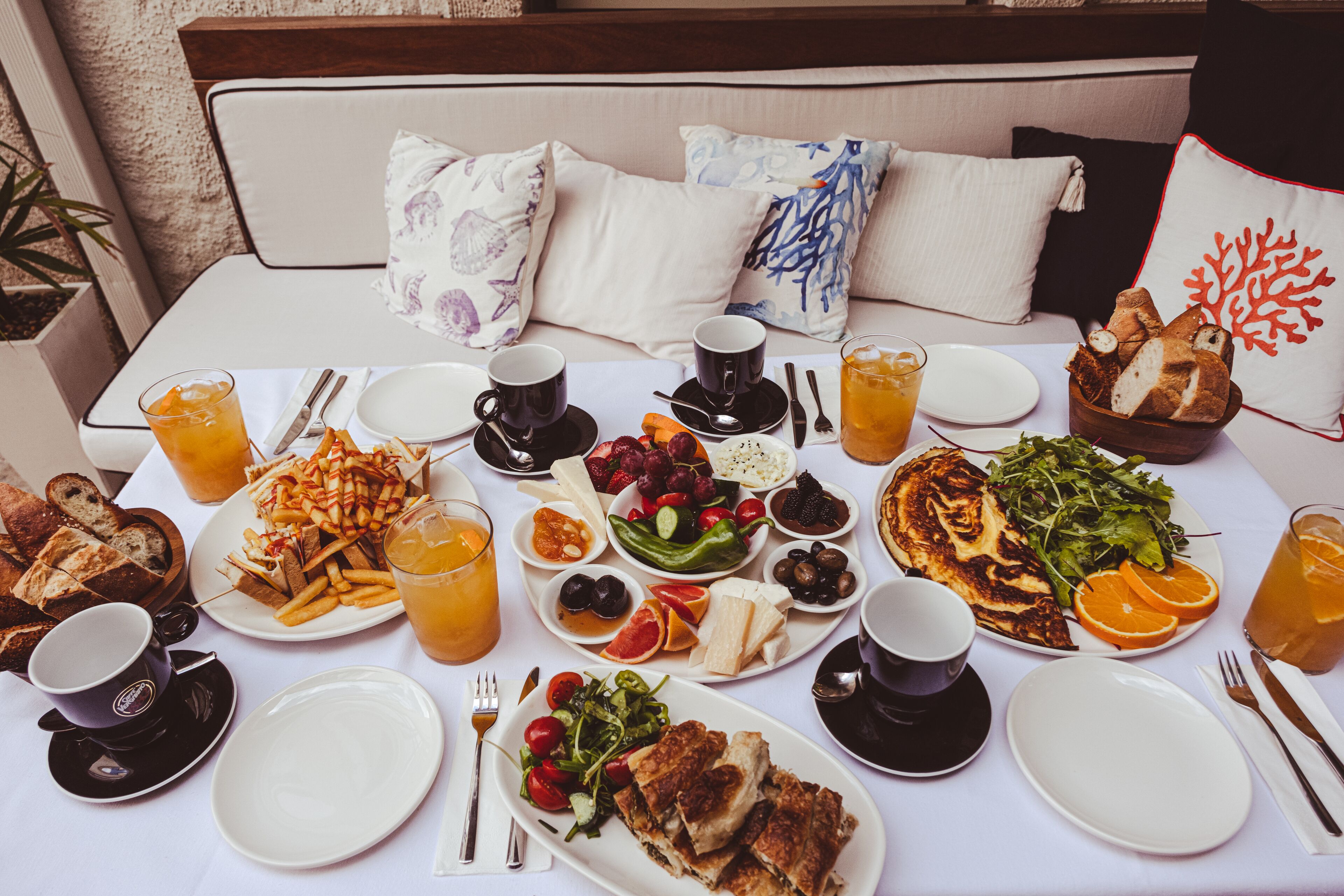 A breakfast setting with a variety of foods and drinks on a table, including bread, fruits, and a salad, with a bed and pillows in the background.