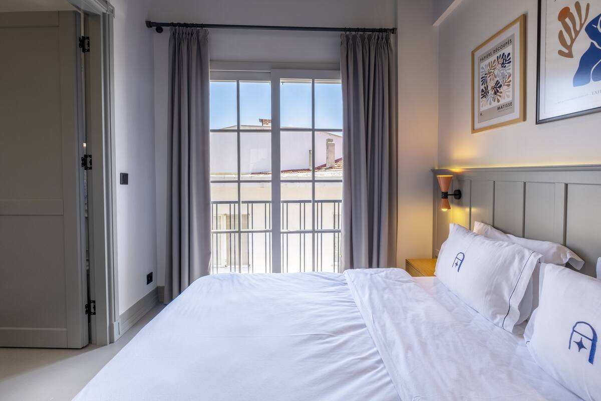 A hotel room with a large bed, a view through a window with curtains, a bedside table with a lamp, and framed artwork on the wall.