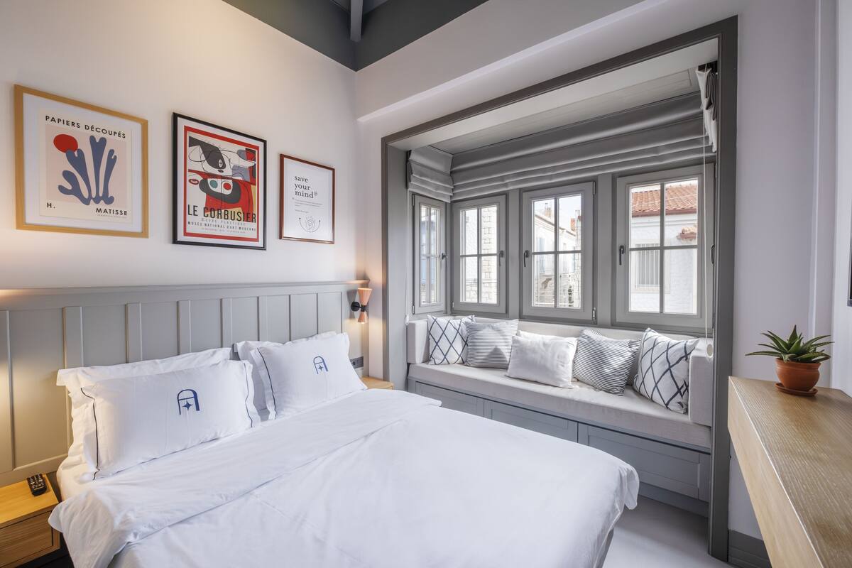 A bedroom with a bed, a window seat with cushions, and three framed artworks on the wall.