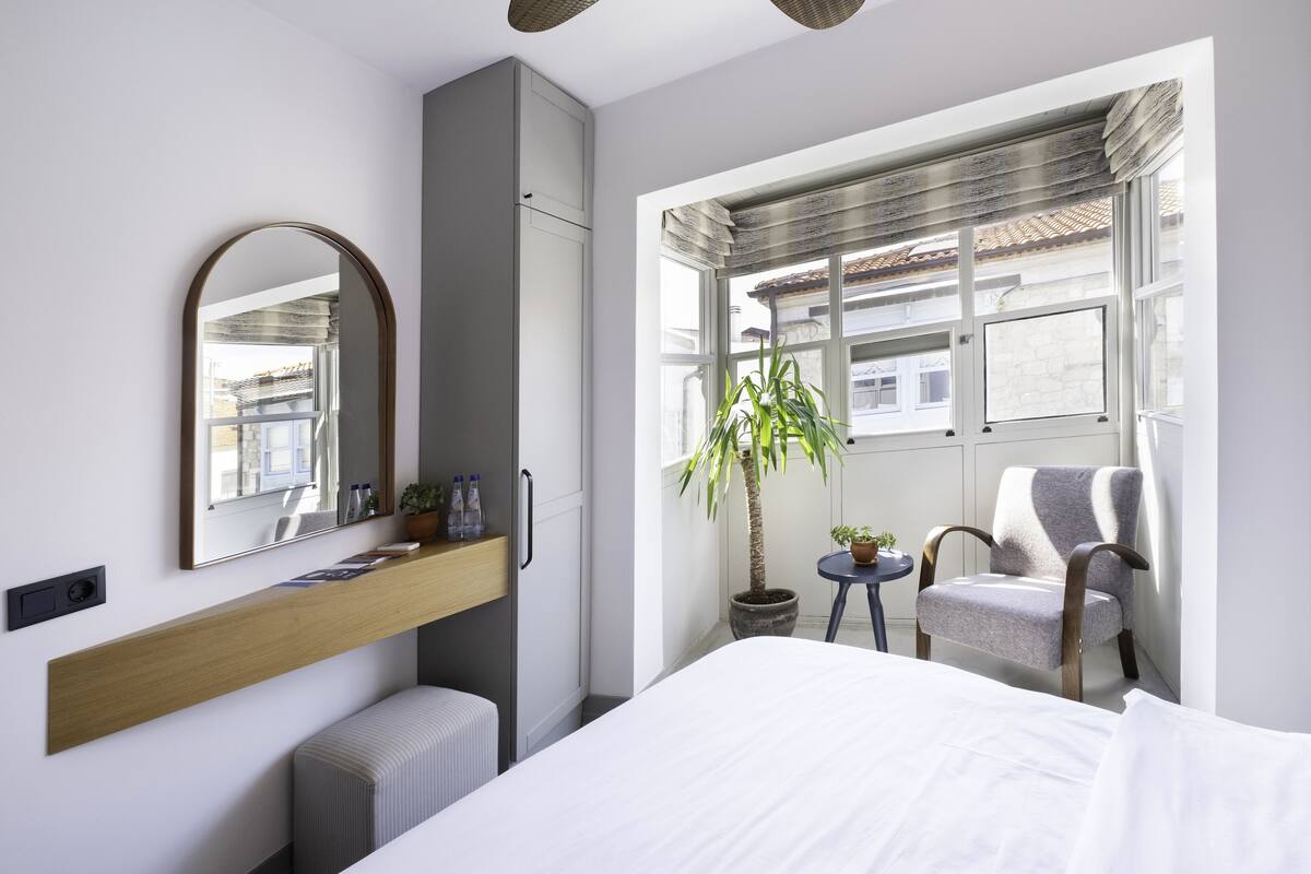 A modern hotel room with a large bed, a wooden bedside table, a mirror, a potted plant, a chair, and a window with a view of buildings.