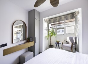 A modern hotel room with a large bed, a wooden bedside table, a mirror, a potted plant, a chair, and a window with a view of buildings. - Alle Alacati (Cesme)