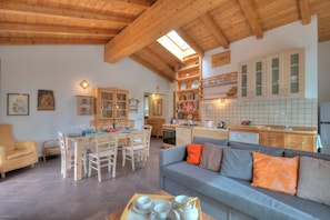 A spacious living area with a wooden ceiling, a grey sofa with orange cushions, a dining table with chairs, and a kitchen area with wooden cabinets and a tiled backsplash. - Casa Tra Borgo e Lago in Lake Tenno (Tenno)
