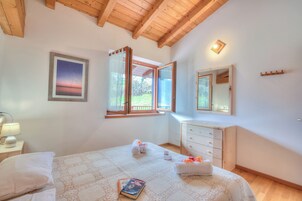 Un dormitorio con cama, mesita de noche con lámpara, cómoda y ventana con vistas a la vegetación.