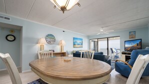 A dining area with a round wooden table, white chairs, and patterned cushions. There are two wall clocks, a ceiling fan, and a television on a stand. - Beautiful Two Bedroom Oceanfront Great Mid Town Location! (Ocean City)
