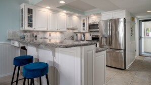 A modern kitchen with white cabinets, a stainless steel refrigerator, a granite countertop, and two blue barstools. - Beautiful Two Bedroom Oceanfront Great Mid Town Location! (Ocean City)