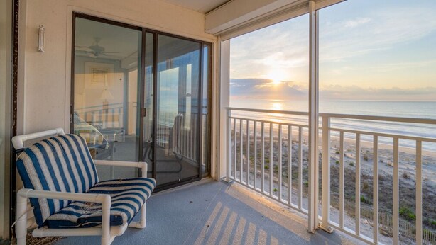 A balcony with a view of the sunset over the ocean, a white chair, and a glass door leading to an interior space. - Beautiful Two Bedroom Oceanfront Great Mid Town Location! (Ocean City)