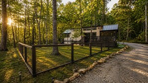 A rustic cabin with a metal fence, surrounded by a forested area with a gravel path leading to it.
