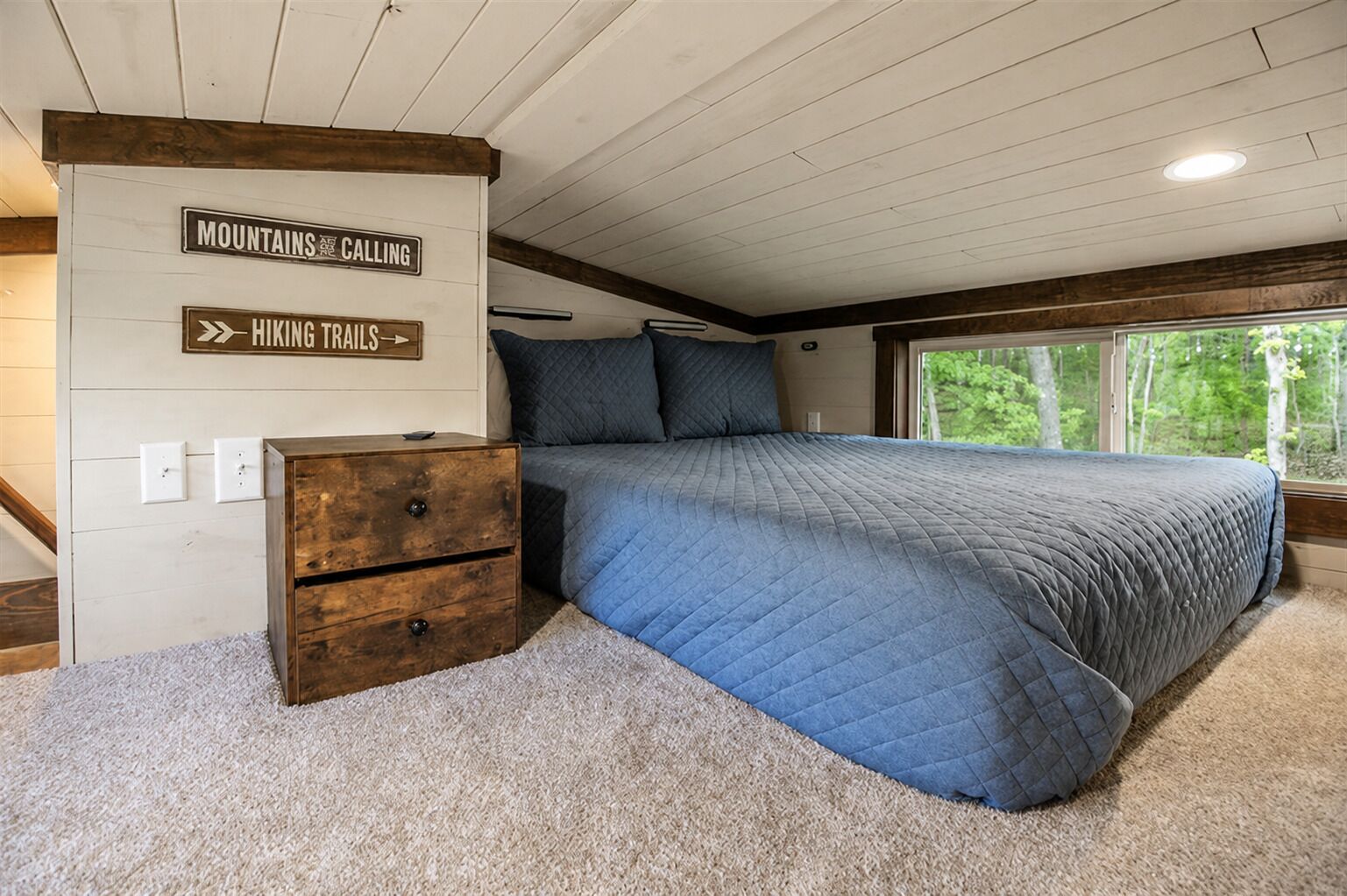 A bedroom with a bed, a dresser, and a window with a view of trees. There are signs on the wall indicating 'Mountains Calling' and 'Hiking Trails.'