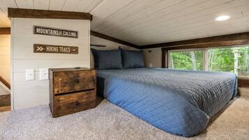 A bedroom with a bed, a dresser, and a window with a view of trees. There are signs on the wall indicating 'Mountains Calling' and 'Hiking Trails.'