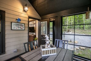 Une terrasse en bois avec une table et des chaises, un tableau encadré et une fenêtre donnant sur les arbres.