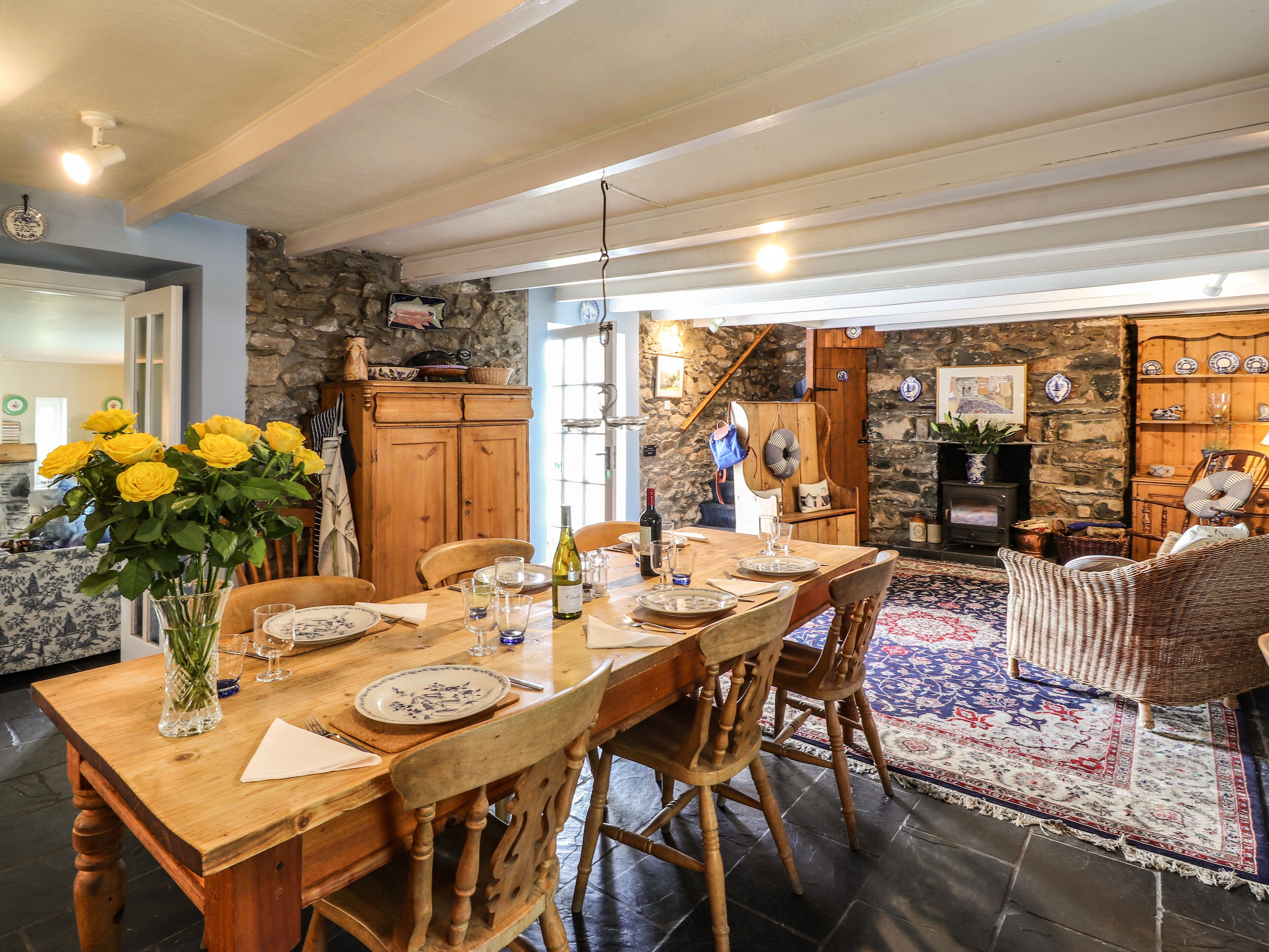 A dining area with a wooden table set for four, surrounded by wooden chairs. A vase with yellow flowers is on the table, and there is a stone wall in the background.