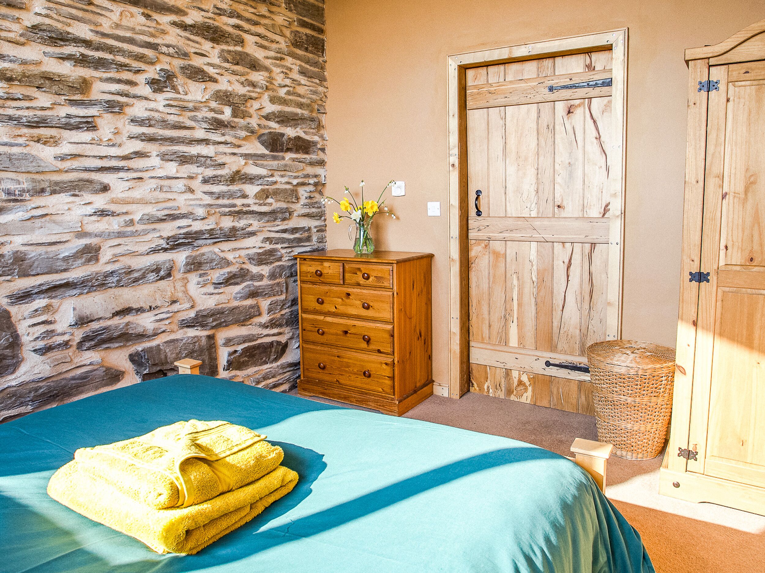 A bedroom with a stone wall, wooden dresser, and a bed with a blue bedspread and yellow towels.