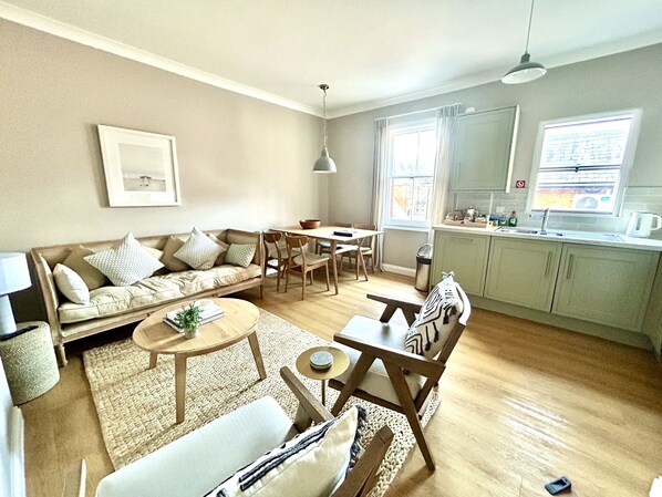 A living room with a sofa, chairs, a coffee table, and a dining area with a table and chairs. There is a kitchen area with cabinets and a window.