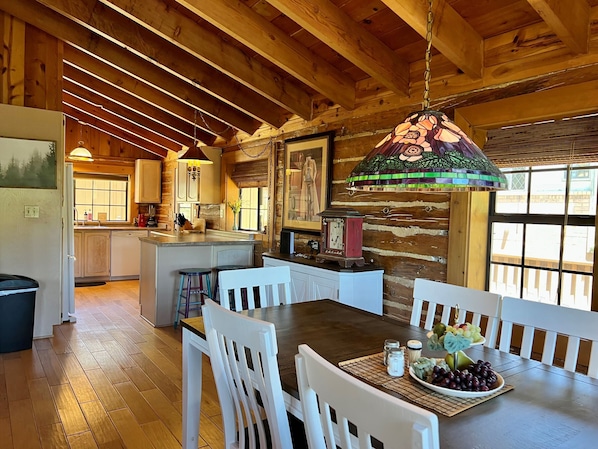 A rustic kitchen with wooden beams, a dining area with white chairs, and a large window.
