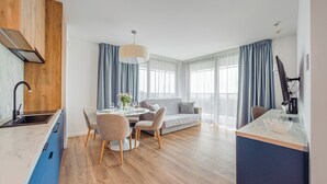 A modern kitchen with a built-in oven, a dining area with a round table and chairs, a sofa with cushions, and a large window with curtains. - Apartamenty Sun & Snow Jantaris (Mielno)