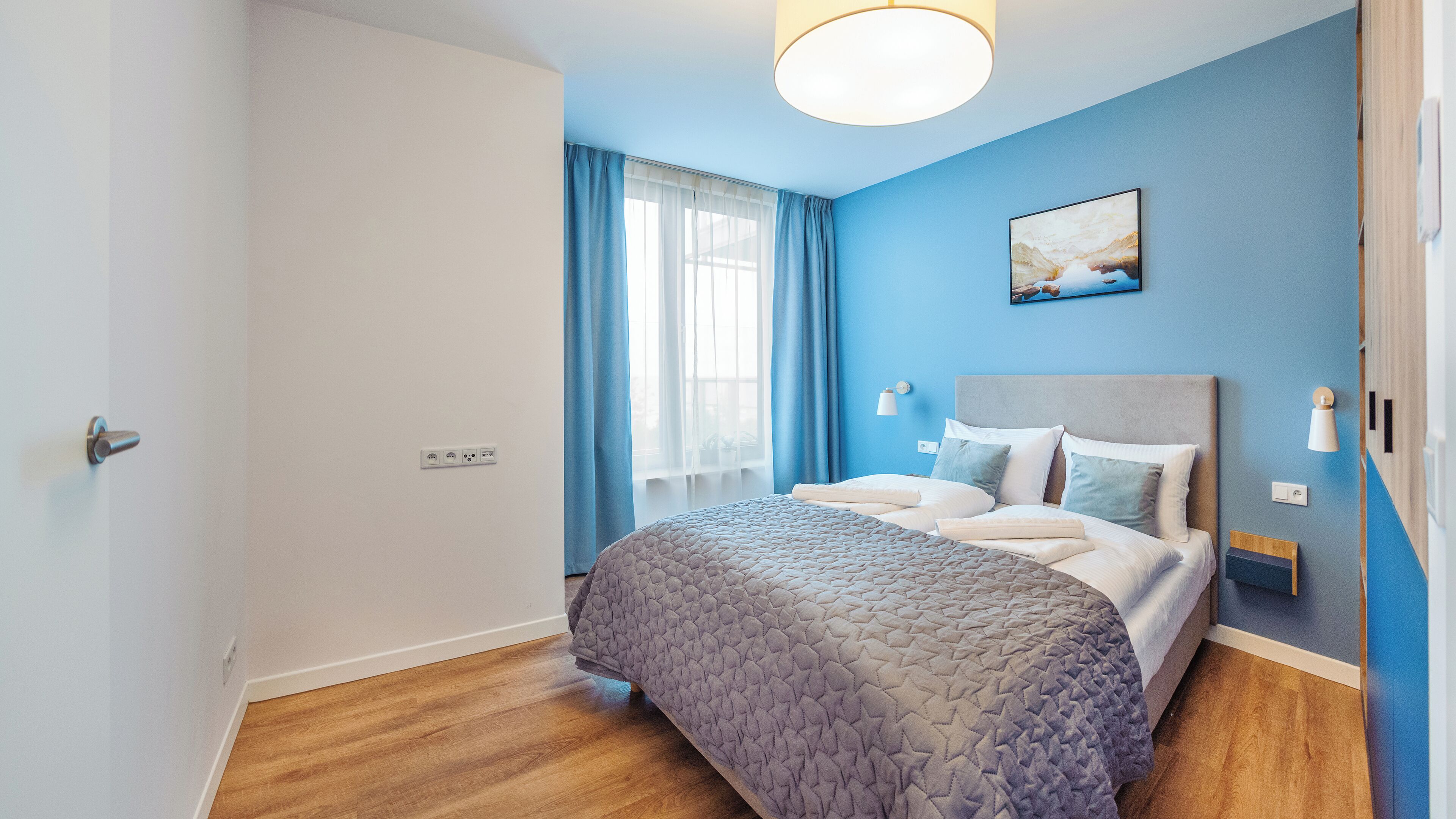 A modern bedroom with a bed, a bedside table, a nightstand, a wall-mounted lamp, and a painting.