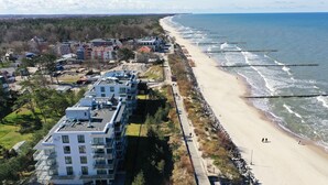 An aerial view of a coastal area with a beach, a row of modern buildings, and a pier extending into the sea. - Apartamenty Sun & Snow Jantaris (Mielno)