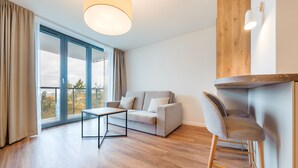 A modern living room with a sofa, a table, and a chair. There is a large window with curtains, a ceiling light, and a wooden accent wall. - Apartamenty Sun & Snow Jantaris (Mielno)