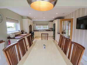 A modern kitchen with a dining area, featuring a large table, wooden chairs, a built-in oven, and a TV mounted on the wall.