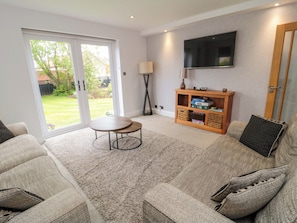 A living room with a sofa, a coffee table, a TV mounted on the wall, and a sliding glass door leading to a garden.