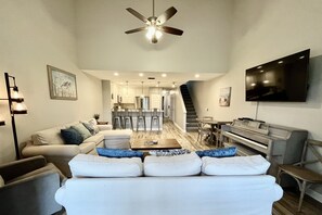 A modern living room with a sofa, coffee table, and a flat-screen TV. There is a dining area with chairs and a piano.