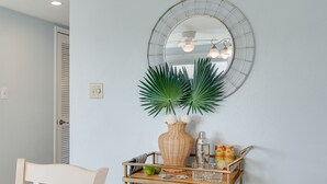 A room with a light blue wall, a round mirror, a wooden chair, a small wooden table with a tray, a wicker vase with green plants, and a ceiling fan. - 300 Ocean Mile: 150 Steps to Beach! Kid Friendly, 2 Pools, Bikes! (Saint George Island)