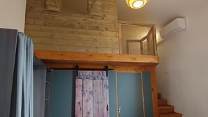 Een slaapkamer met een houten plafond, een trap, een bed, een raam met gordijnen en een airconditioningsunit.