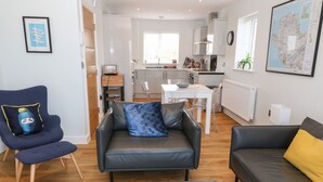 A modern living room with a grey sofa, a blue armchair, and a yellow cushion. There is a dining table with chairs, a kitchen area, and a large map on the wall. - Beach Apartment 9 (Holyhead)