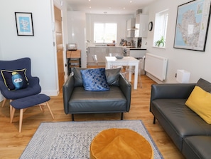 A modern living room with a grey sofa, a blue armchair, and a yellow cushion. There is a dining table with chairs, a kitchen area, and a large map on the wall. - Beach Apartment 9 (Holyhead)