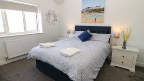 A bedroom with a large bed, a bedside table, a nightstand, a window, a radiator, and a painting of a beach scene. - Beach Apartment 9 (Holyhead)