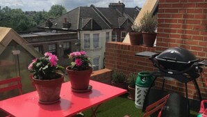 A rooftop terrace with a red table and chairs, a barbecue grill, and potted plants. - Unique 2 Bedroom Penthouse Apartment in Kensal Rise (London)