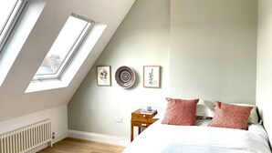 A bedroom with a sloped ceiling, skylights, a bed with red and white bedding, a bedside table with a book, and framed botanical prints on the wall. - Unique 2 Bedroom Penthouse Apartment in Kensal Rise (London)