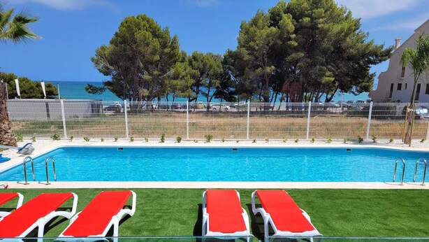 A swimming pool with red lounge chairs and a view of the sea. - Azahar Beach Apartments (Alcala de Xivert)