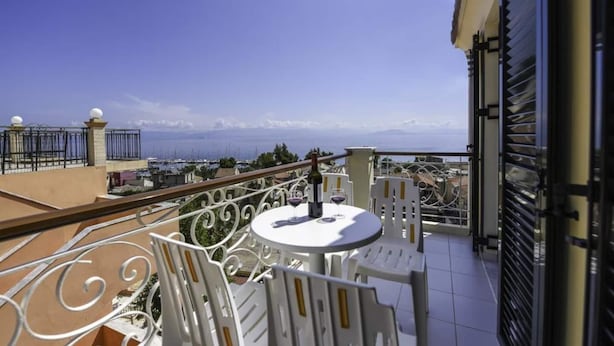 Ein Balkon mit weißem Tisch und Stühlen, der Blick ins Küstenlandschaft mit Gebäuden und Bergen in der Ferne gewährt.