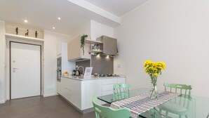 Una cocina moderna con armarios blancos, estufa integrada y una pequeña isla. Hay una mesa de comedor de vidrio con sillas verdes y un jarrón con flores amarillas.