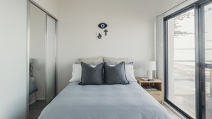 A modern bedroom with a large bed, a nightstand, a wall-mounted lamp, and a view of the outdoors through a large window. - Sandy Point Beach House - On The Waterfront.  Mid Week Stay Special Is Now On. (Corlette)